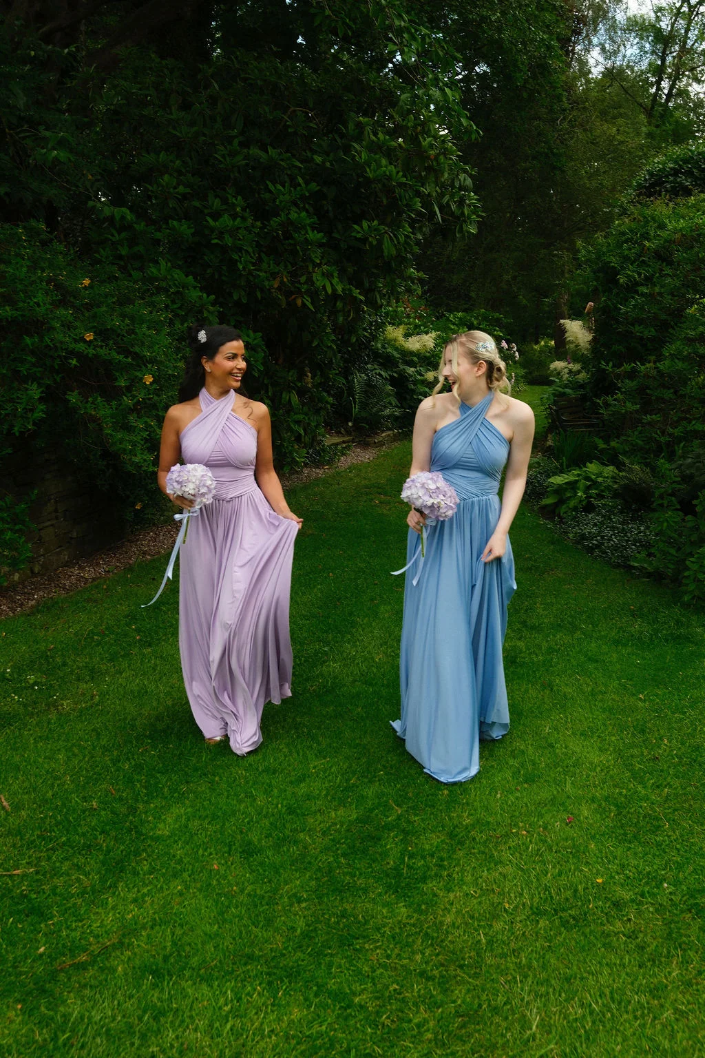 Two bridesmaids in mismatched halterneck dresses of dusty pink and pastel blue walk through a lush green garden while holding matching bouquets and laughing together.