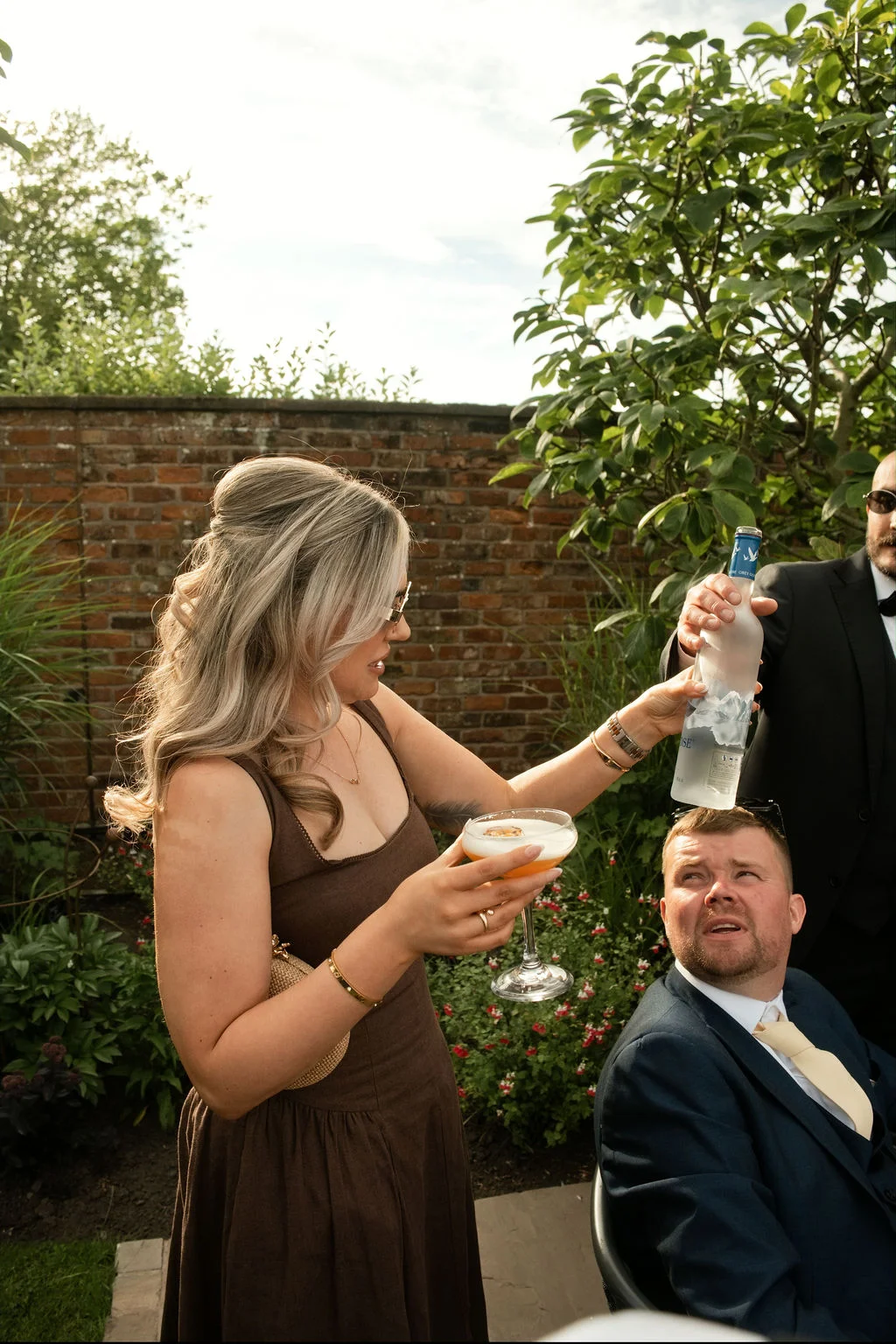 A wedding guest pours from a bottle above a seated man's head during an outdoor drinks reception in front of a brick wall.