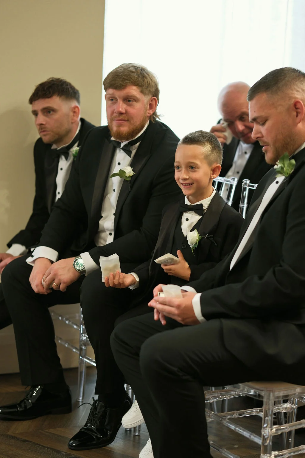 A group of groomsmen and a page boy in black tuxedos sit together indoors, holding small packets while smiling during the wedding ceremony.