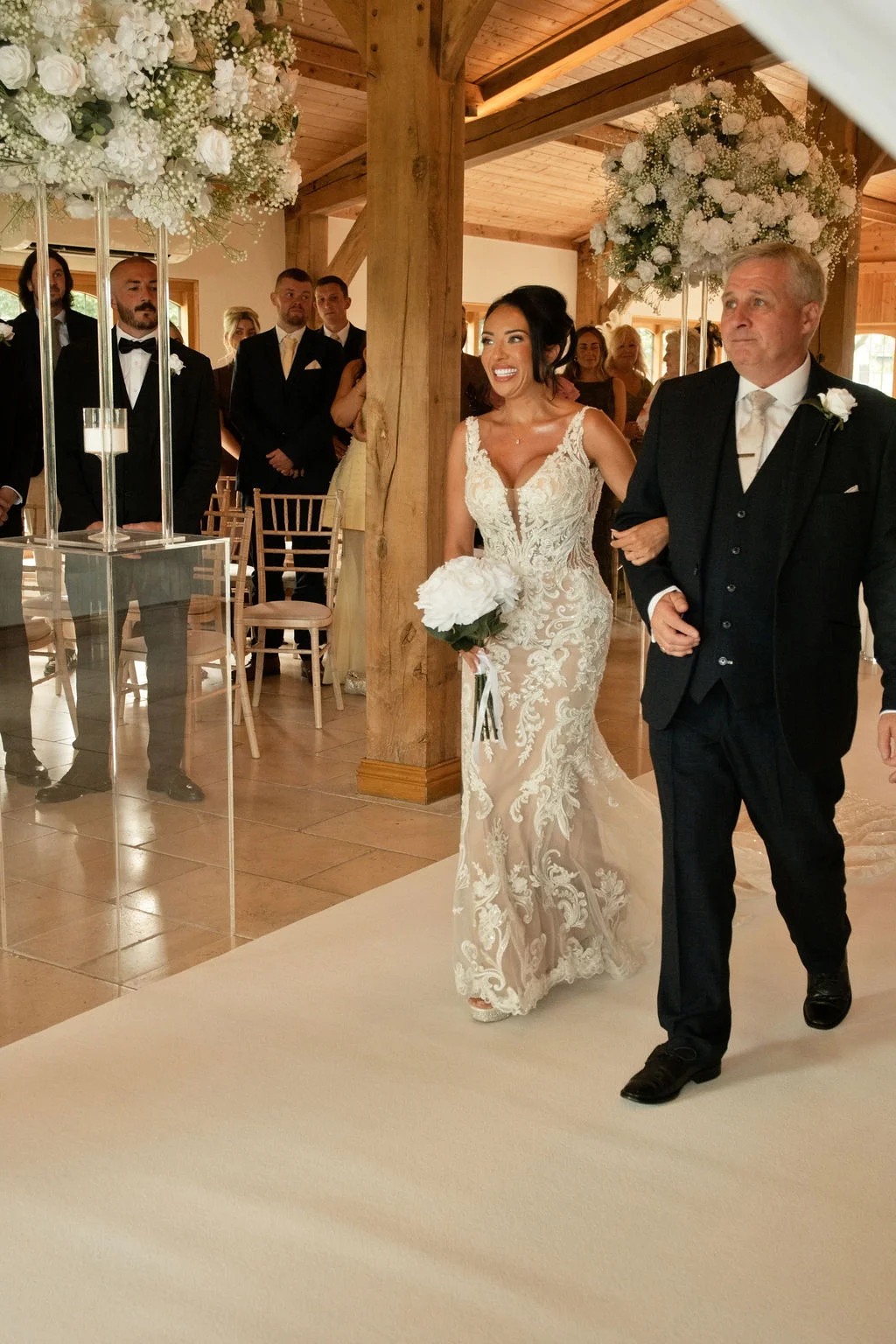 A joyful bride walks down the white aisle of a rustic oak-framed barn on the arm of her father, holding a white bouquet while wedding guests watch from chairs.
