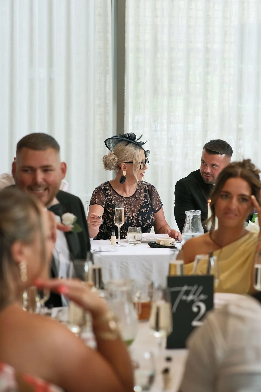 Guests enjoy lively conversation and drinks during the wedding breakfast seated at elegantly decorated white tables indoors.
