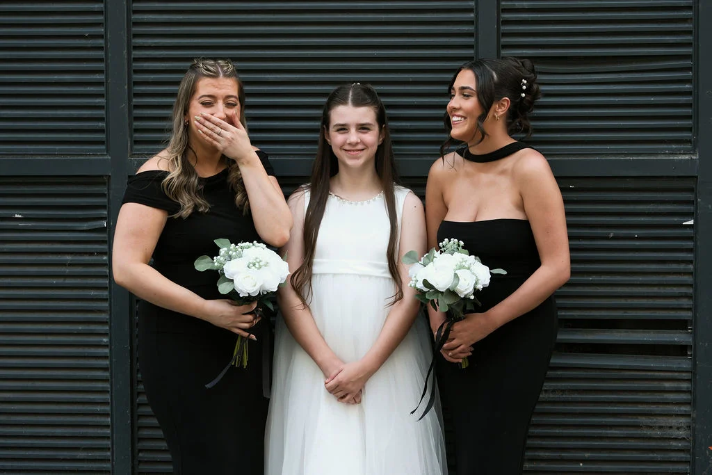 Two bridesmaids in black dresses and a junior bridesmaid in a white dress stand against a dark slatted wall, holding white bouquets while laughing.