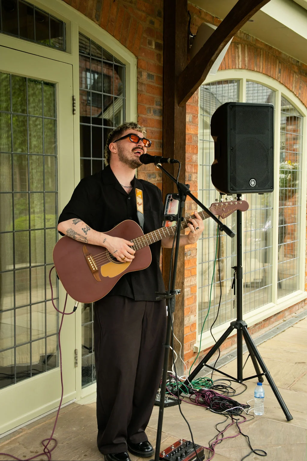 A stylish male singer with sunglasses and tattoos performs live with an acoustic guitar outdoors in front of arched brick and glass windows.