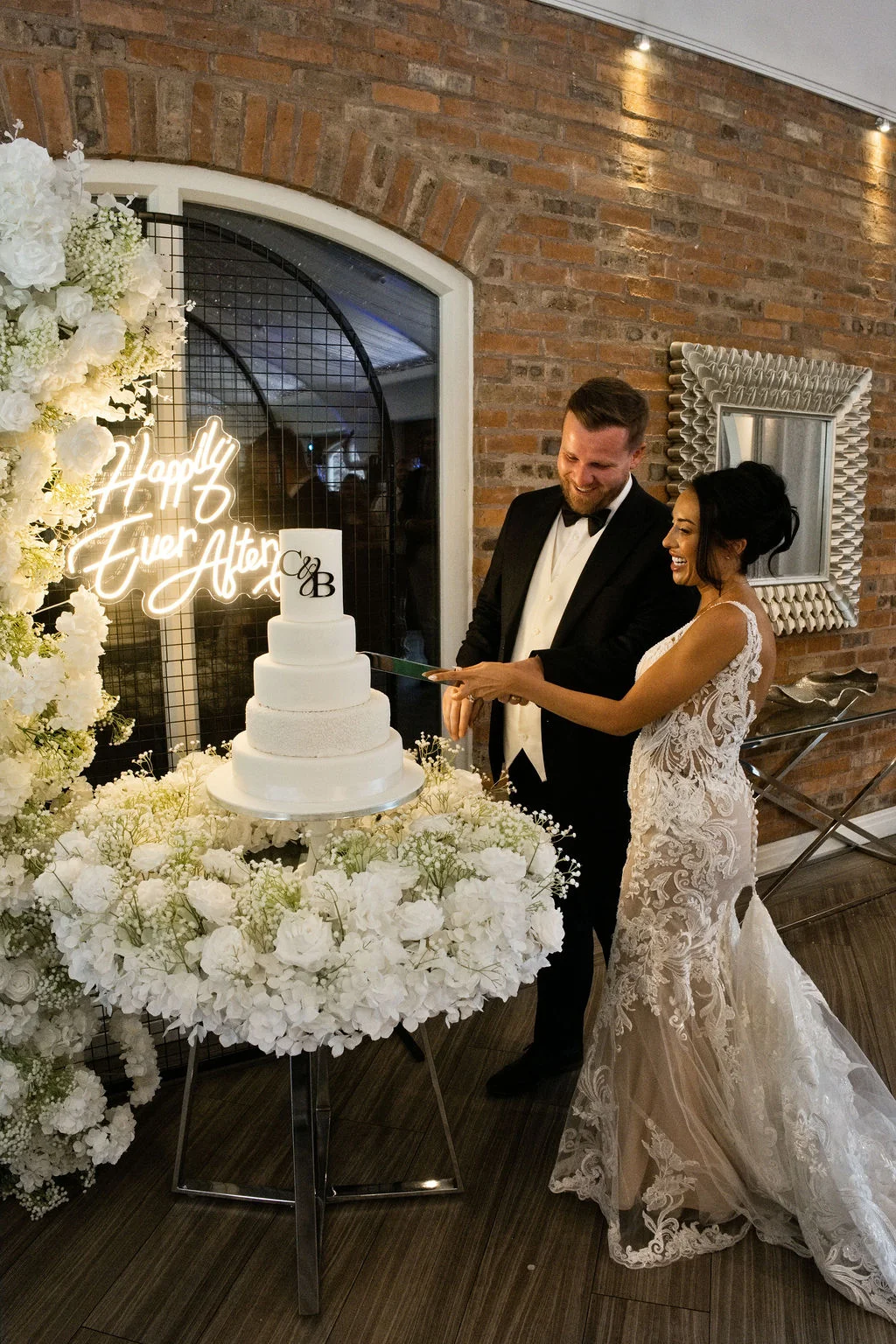 A smiling bride and groom cut a multi-tiered white wedding cake against an exposed brick wall featuring a glowing neon sign and abundant floral arrangements.