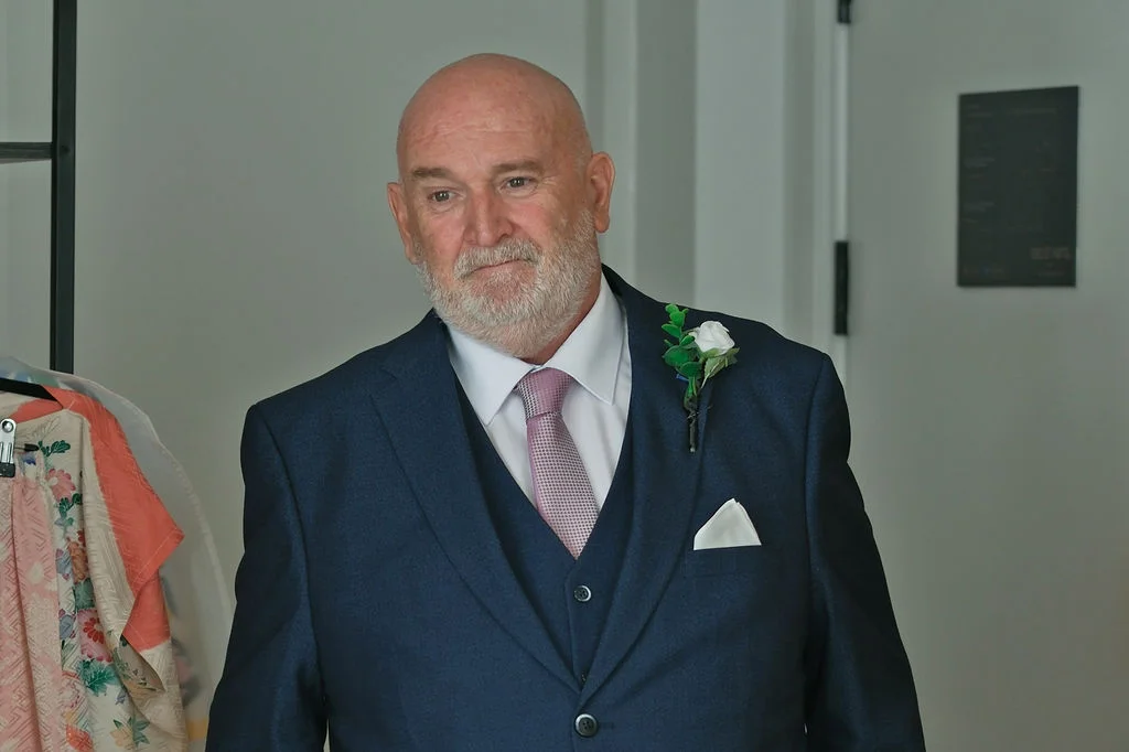 A mature man in a smart navy three-piece suit wearing a buttonhole with a white rose looks thoughtfully ahead.