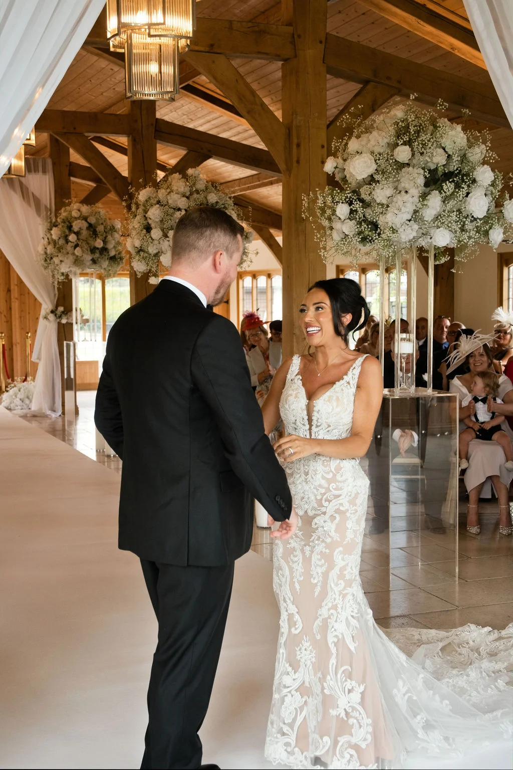 A joyful newlywed couple stand at the aisle inside a sunlit oak-framed barn featuring tall floral arrangements and seated guests.