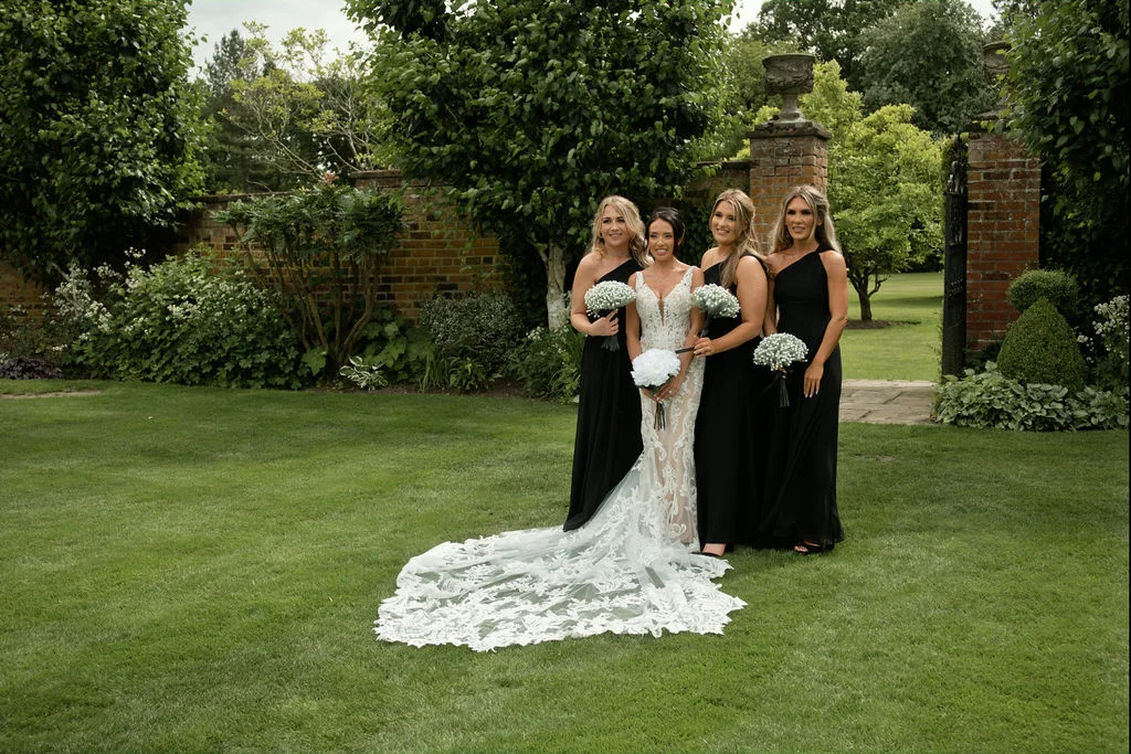 A stunning bride in an intricate lace gown with a dramatic train poses on a manicured lawn with four bridesmaids wearing elegant one-shoulder black dresses.