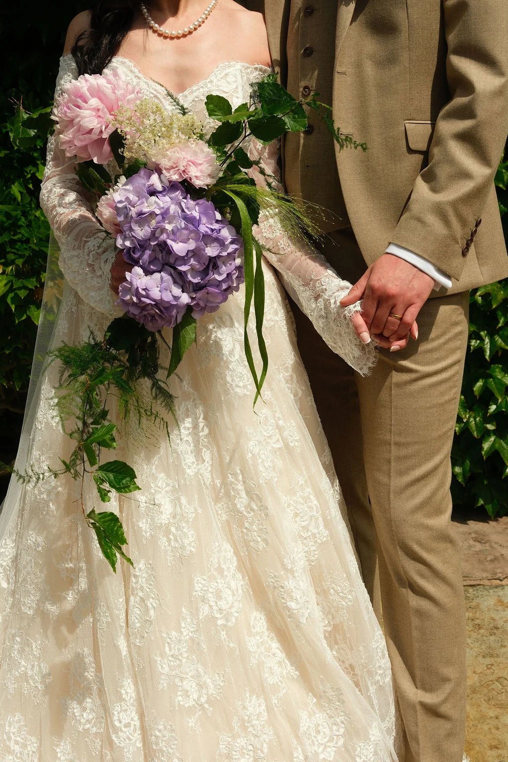 A bride in a lace off-the-shoulder gown holding a large trailing bouquet of hydrangeas and peonies walks hand-in-hand with a groom wearing a tweed suit.