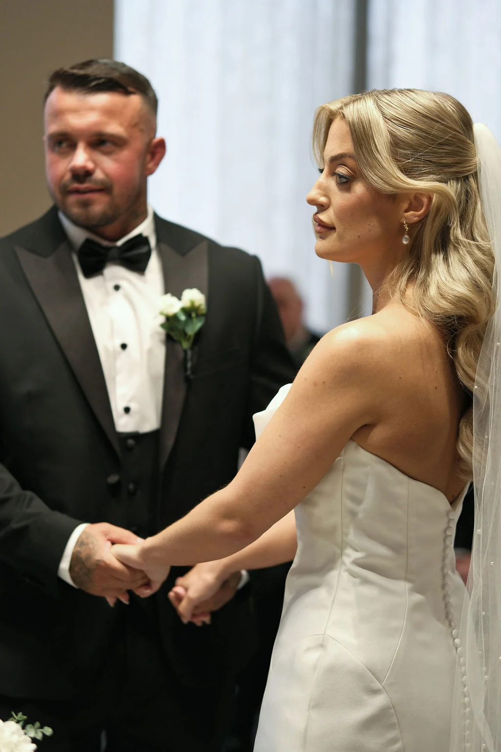 The bride and groom hold hands during their wedding ceremony, with the bride looking thoughtfully aside in her strapless gown and the groom standing in a classic black tuxedo.