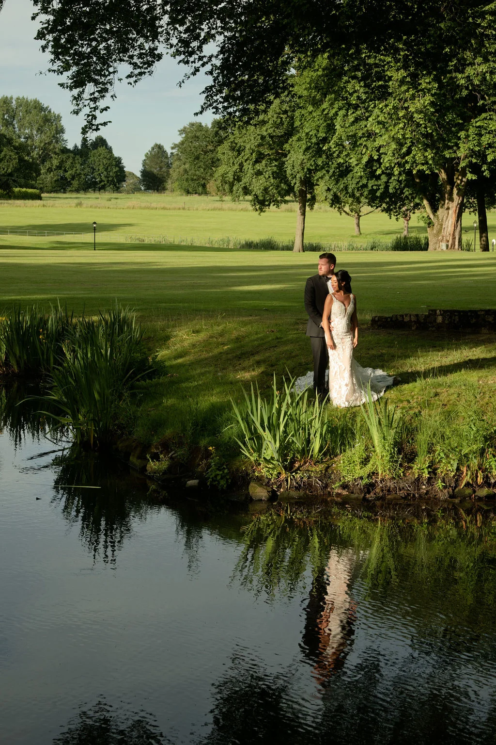 A newlywed couple stands by a calm lake in the late afternoon sun, their reflections captured in the water against a sprawling estate backdrop.