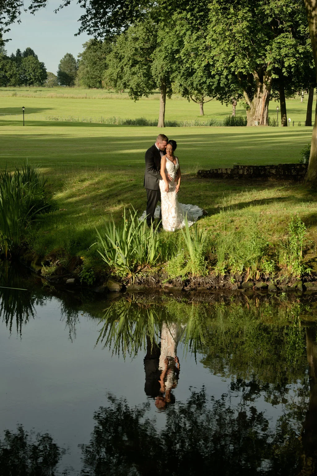 A newlywed couple embraces tenderly near a calm lake during golden hour, their reflection mirrored clearly in the water.
