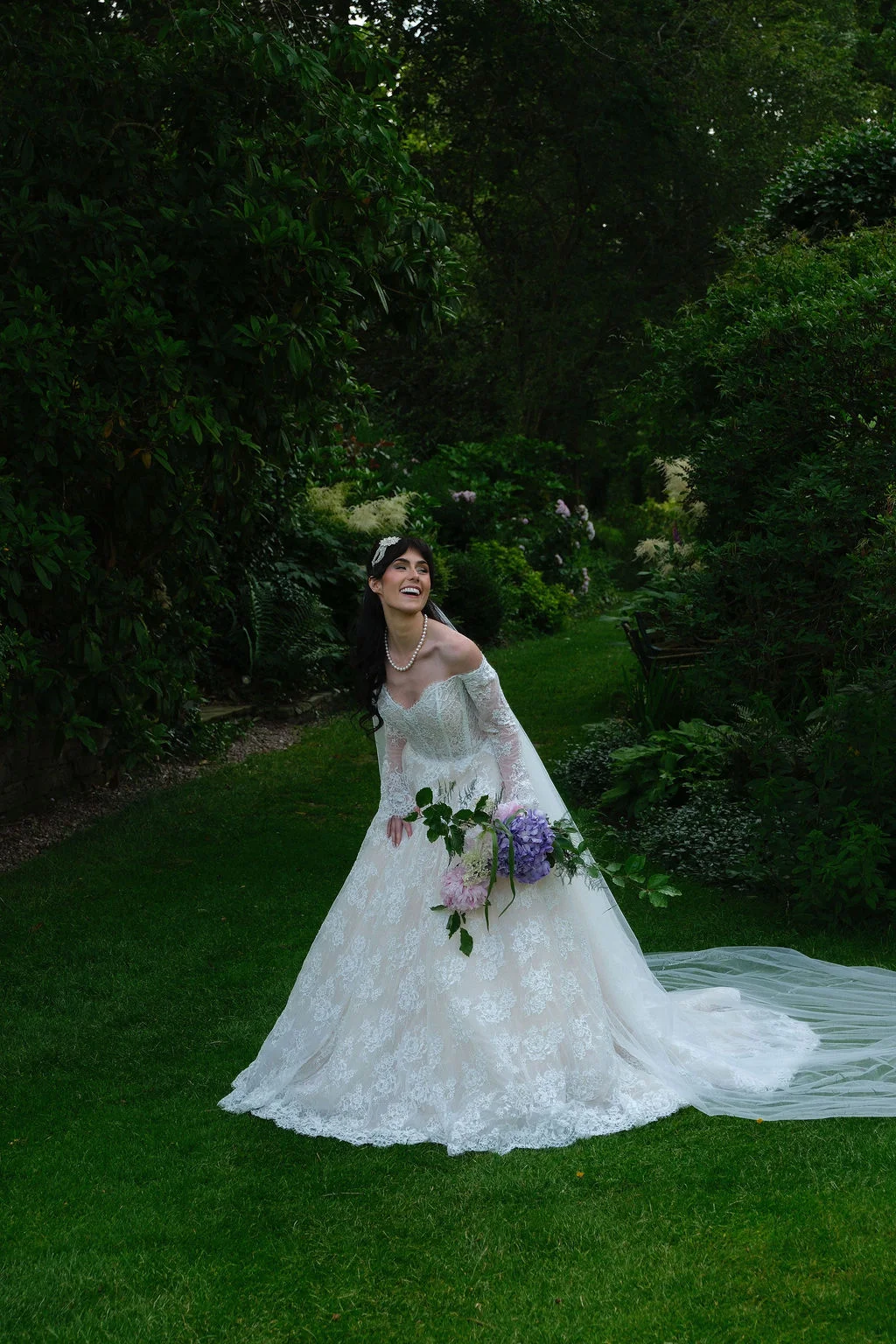A joyful bride in a lace off-the-shoulder gown and long veil laughs while holding a floral bouquet in a lush green woodland garden.