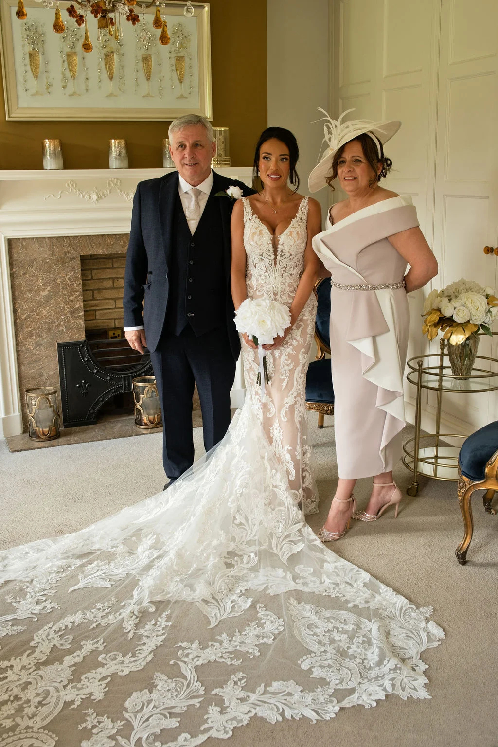 A bride in a plunging lace gown with an elaborate train stands proudly between her parents inside an elegant room with a fireplace.