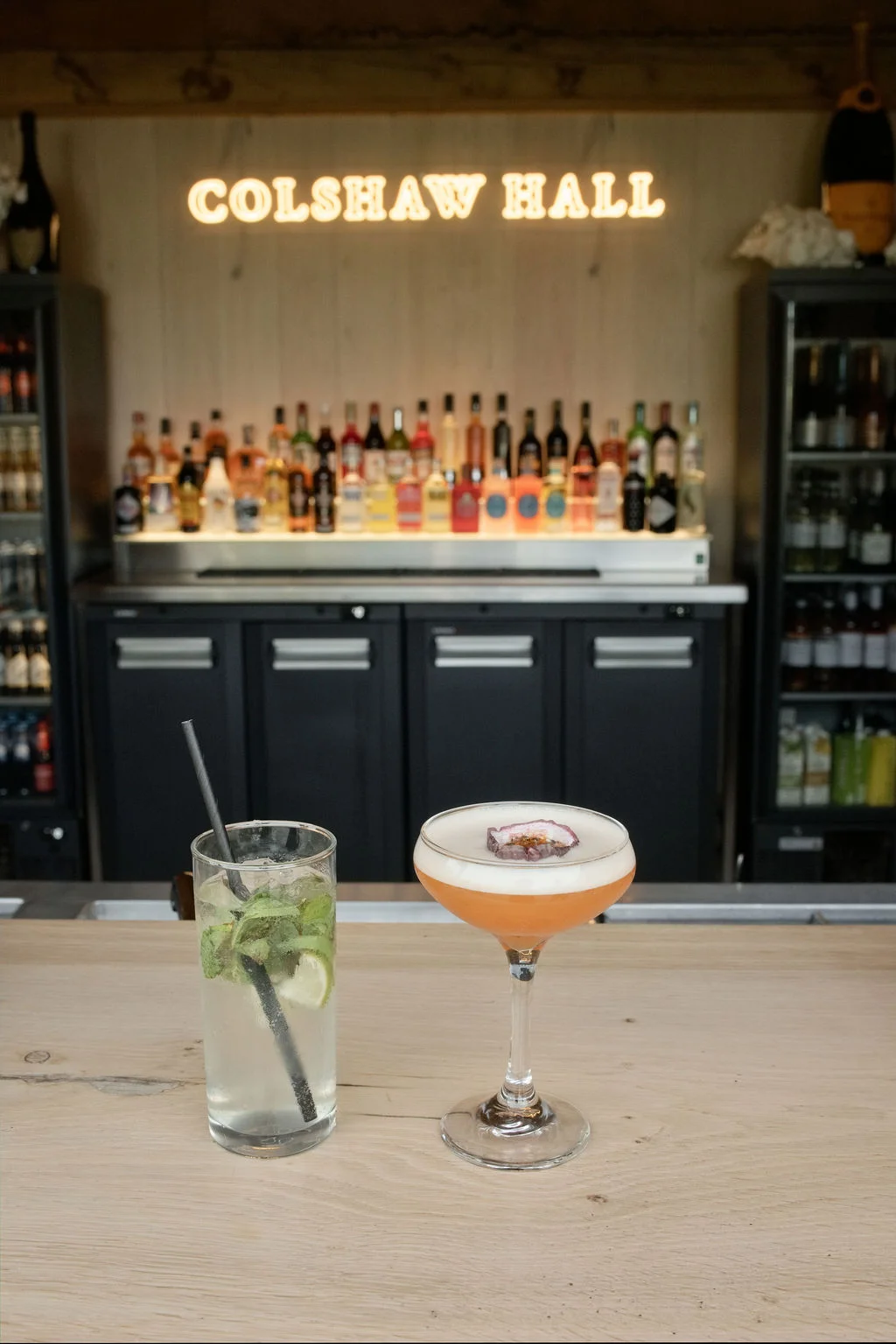 Two freshly prepared wedding cocktails, including a mojito and a pornstar martini, rest on a wooden bar counter beneath an illuminated neon sign.