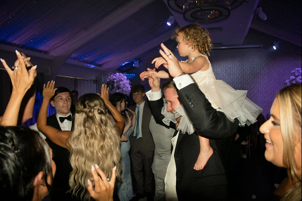 A young wedding guest sits on a man's shoulders on a packed, lively dance floor under purple lighting and ceiling draping.