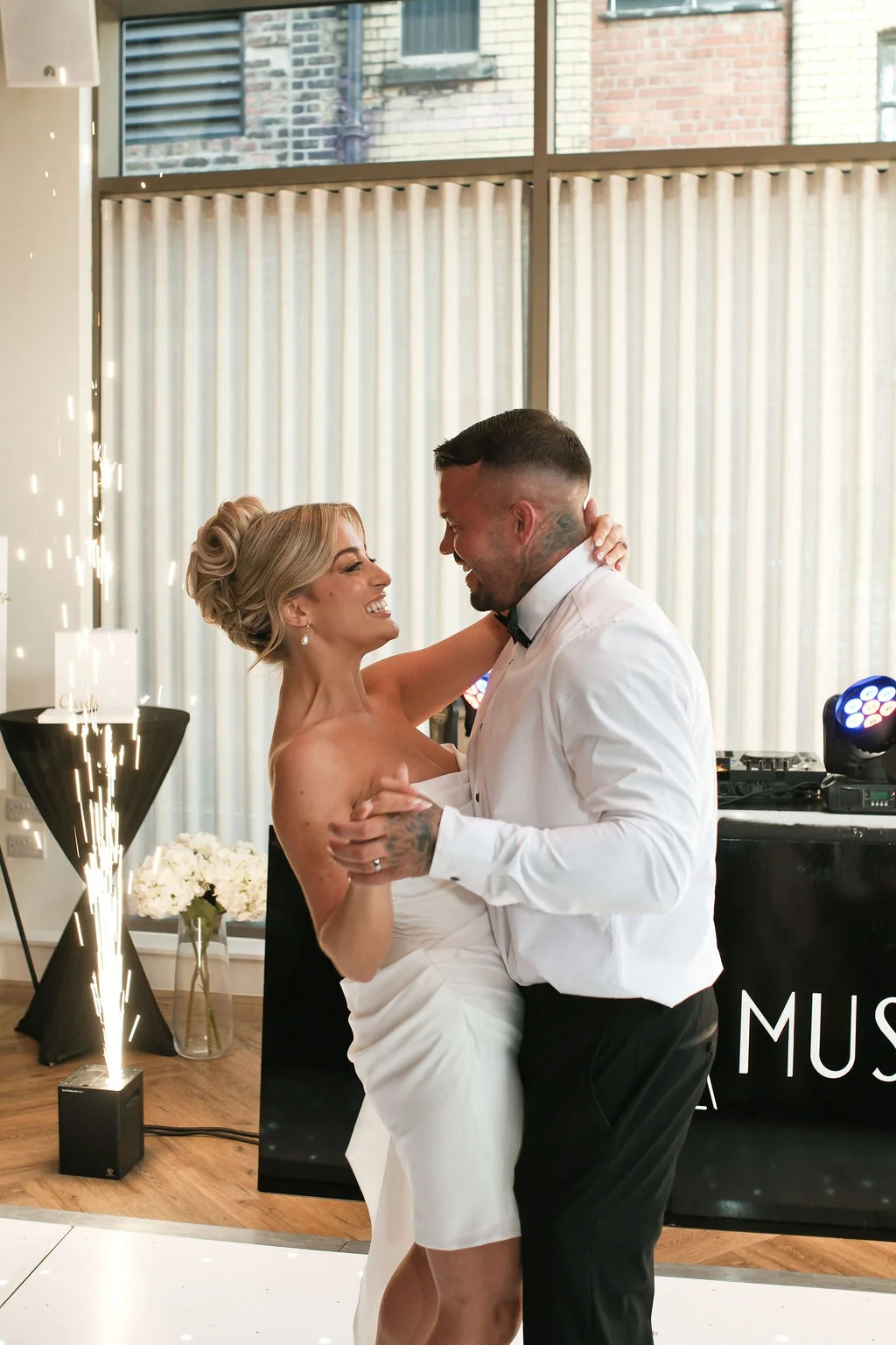 A newlywed couple shares a joyful first dance on an illuminated dance floor while indoor sparklers fountain nearby.