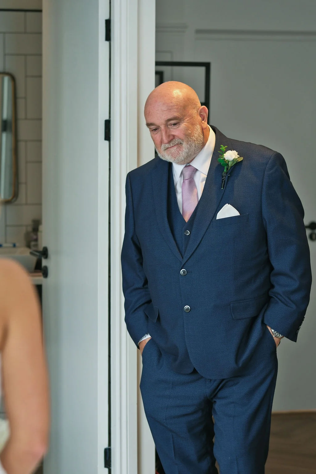 An emotional father of the bride wearing a smart navy three-piece suit smiles down proudly during morning wedding preparations.