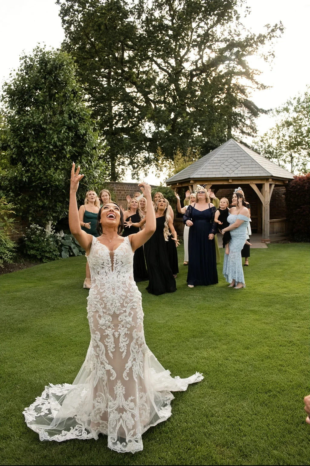 A joyful bride in a lace fit-and-flare gown throws her bouquet outdoors on a green lawn while wedding guests watch with anticipation near a wooden gazebo.