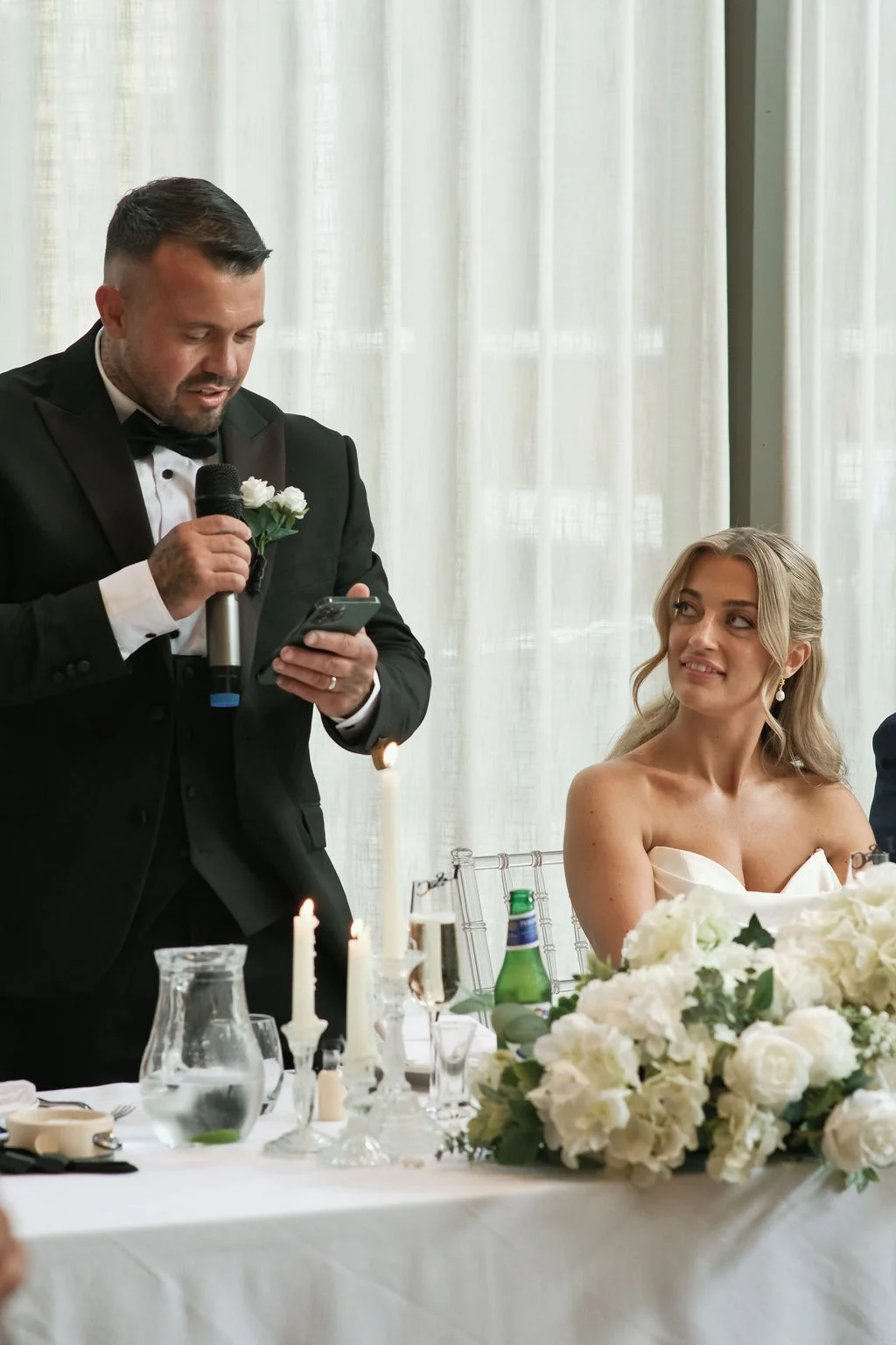 A groom in a black tuxedo stands delivering a speech while holding a microphone and smartphone, looking down lovingly at his smiling bride seated beside him at a beautifully decorated top table adorned with white florals and candlelight.