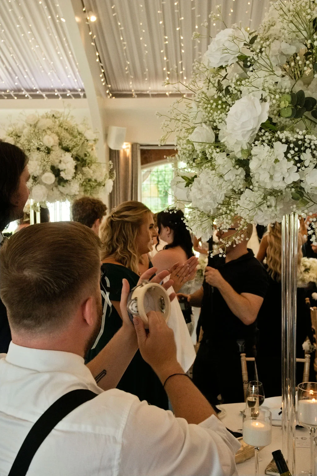 An over-the-shoulder shot of a guest holding a small tambourine during a lively wedding reception featuring tall white floral centerpieces and a fairy light canopy.