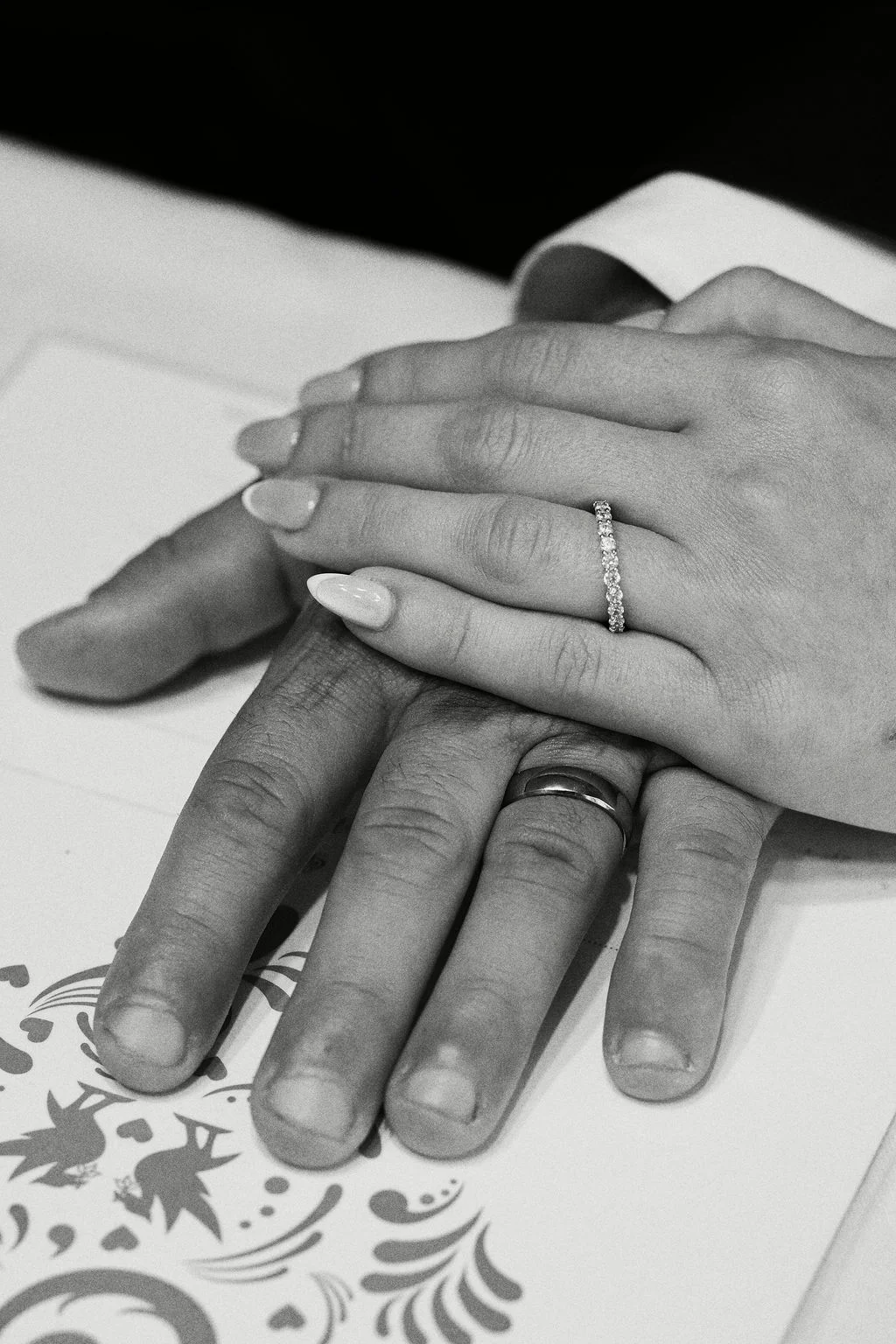 A close-up black and white photograph captures the newly married couple's hands resting together, showcasing their sparkling diamond and plain metal wedding bands.