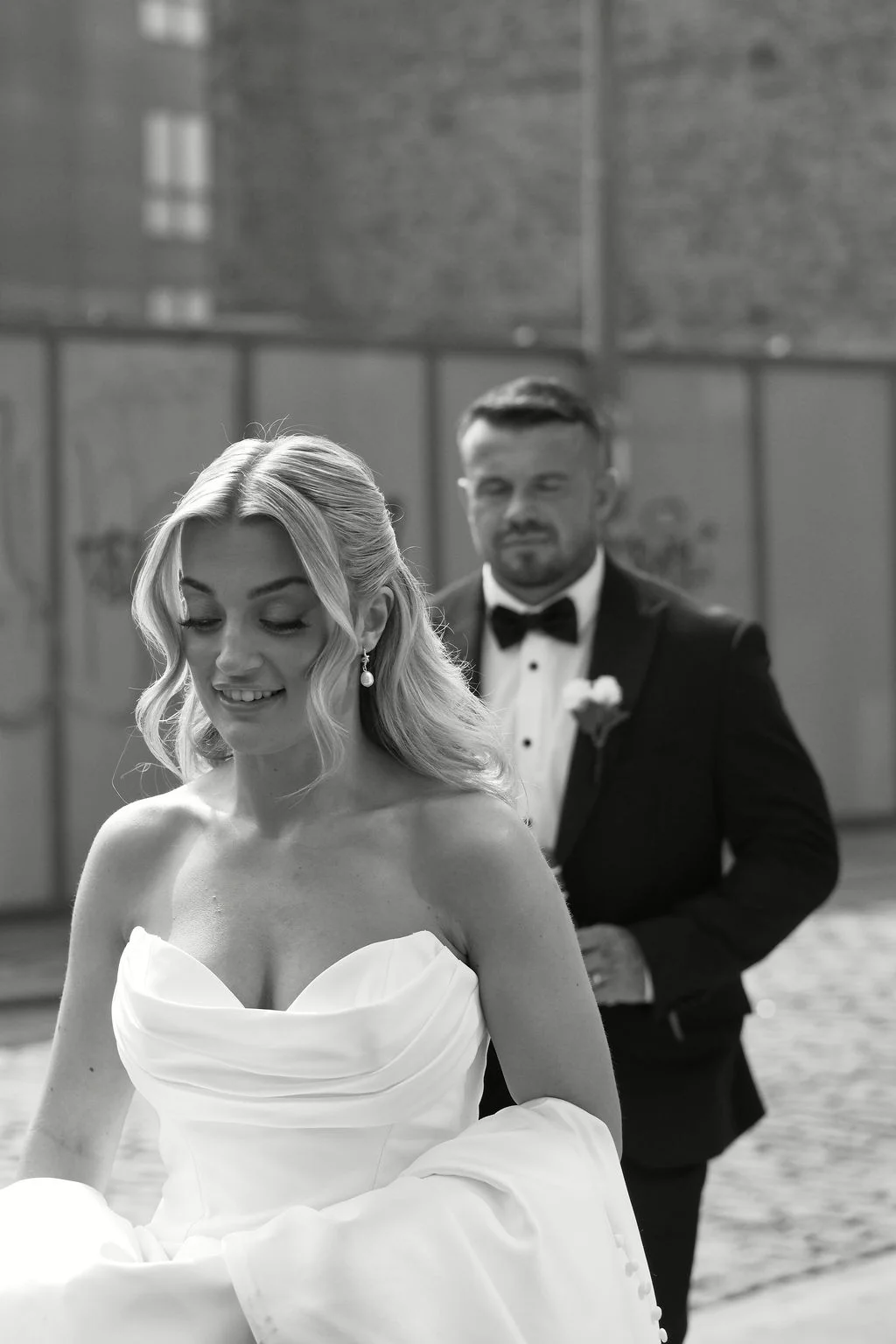 A stunning black and white shot captures a bride in a strapless satin gown walking outdoors, with her groom in a black tuxedo following closely behind on a cobbled courtyard.
