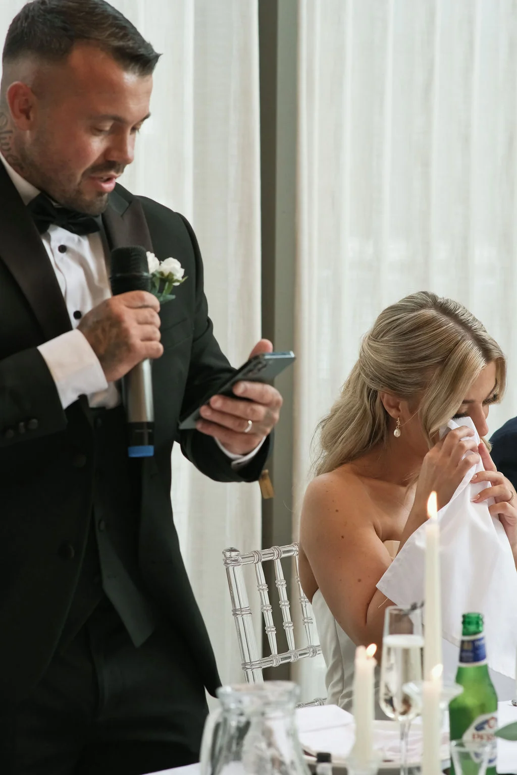 A groom delivers a heartfelt speech holding a microphone and his phone, while the bride wipes away tears with a white napkin at the top table.