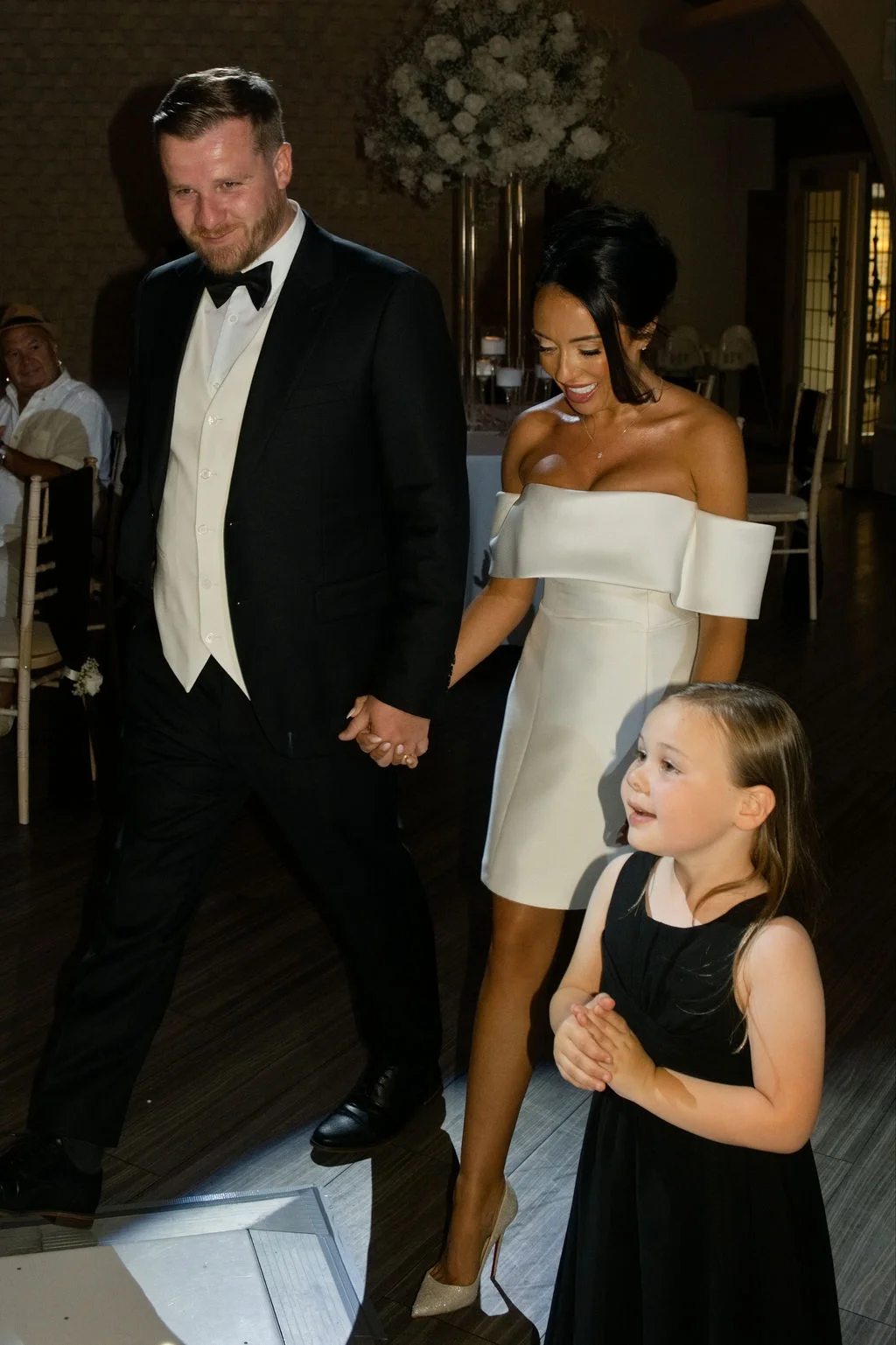 A newlywed couple holding hands as they make their entrance into the reception room, accompanied by a young flower girl.