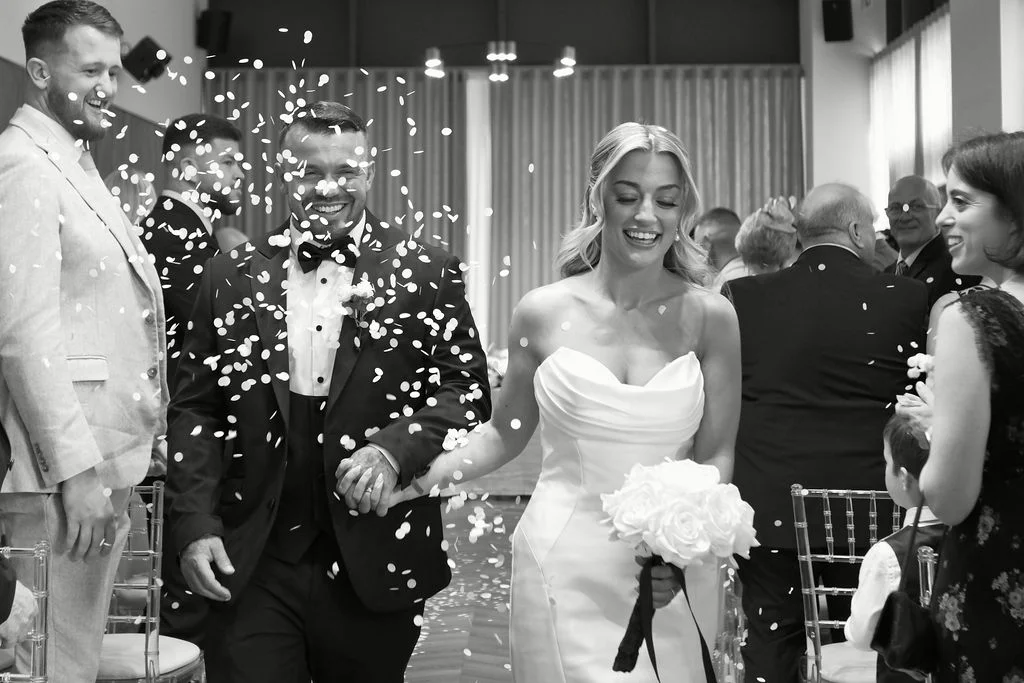A joyful newlywed couple walk hand-in-hand down the aisle amidst a shower of white confetti, smiling brightly as guests cheer.
