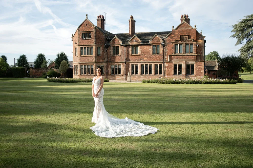 A bride in an intricate lace wedding dress with a dramatic train stands on a sweeping lawn in front of a grand red brick manor house.