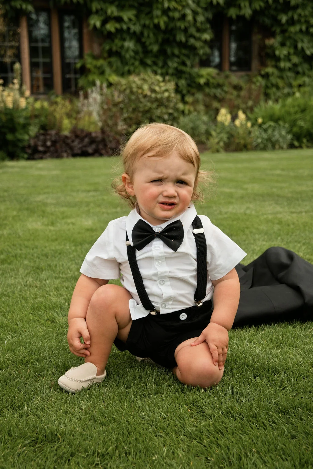 A young page boy wearing a white shirt, black shorts, braces, and a bow tie sits on a green lawn outdoors.