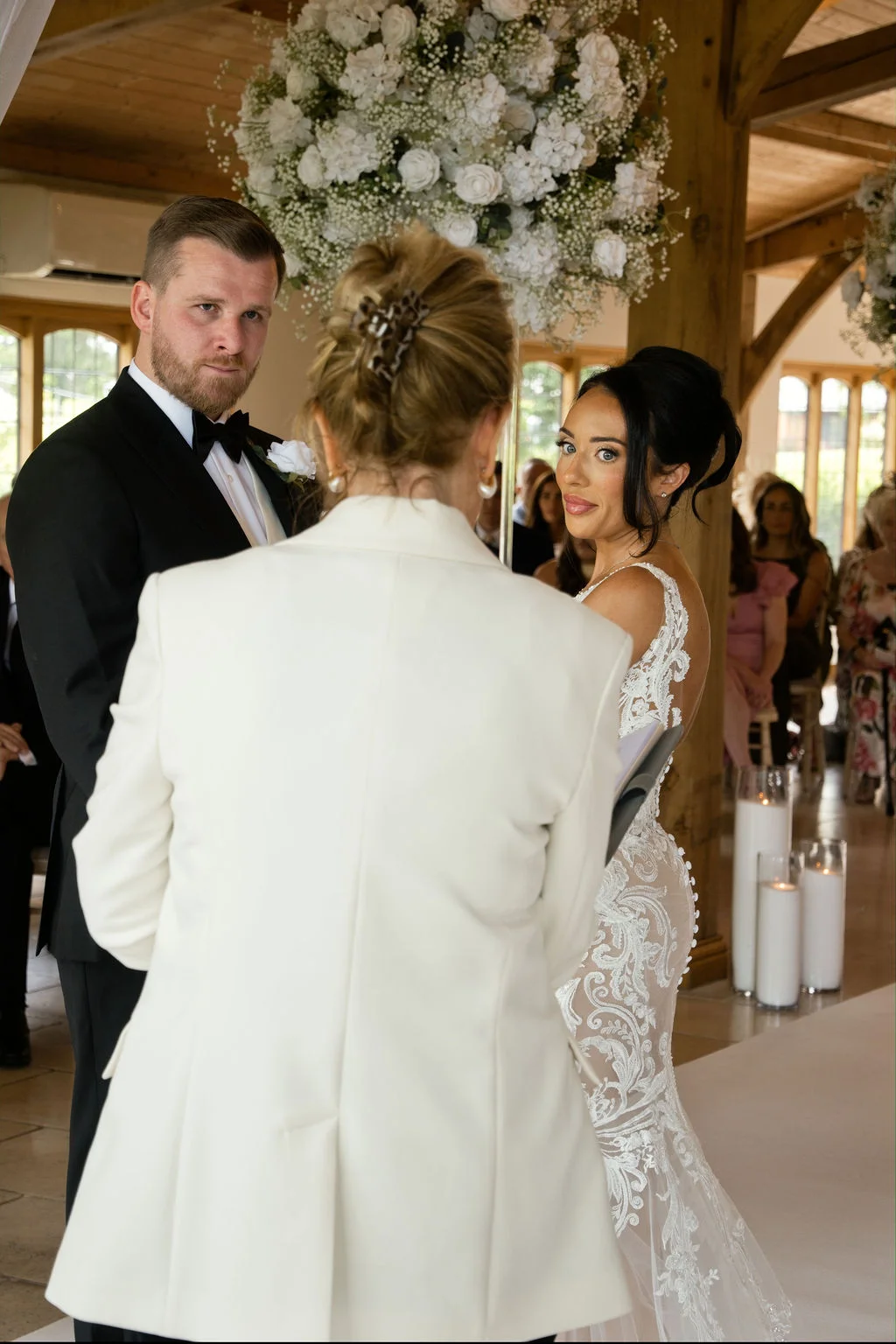 A bride in a lace mermaid gown and groom in a black tuxedo stand before a celebrant during an indoor barn ceremony beneath large floral arrangements.