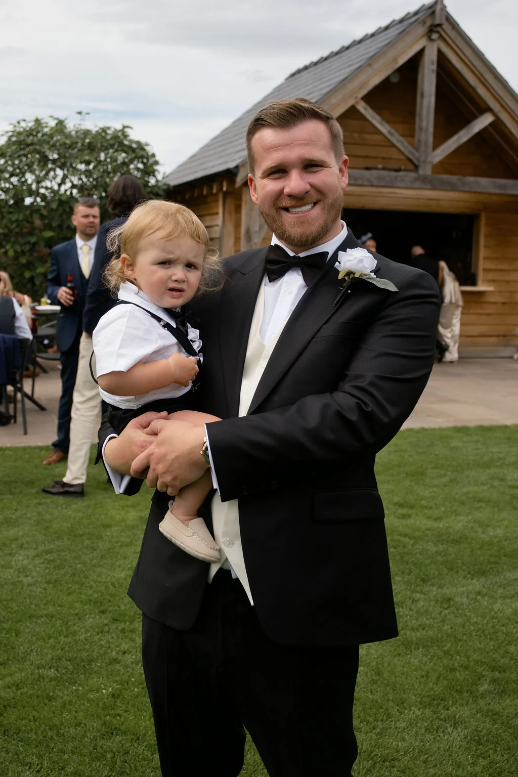 A smiling groom wearing a black tuxedo holds a crying toddler page boy outside a rustic wooden barn venue.