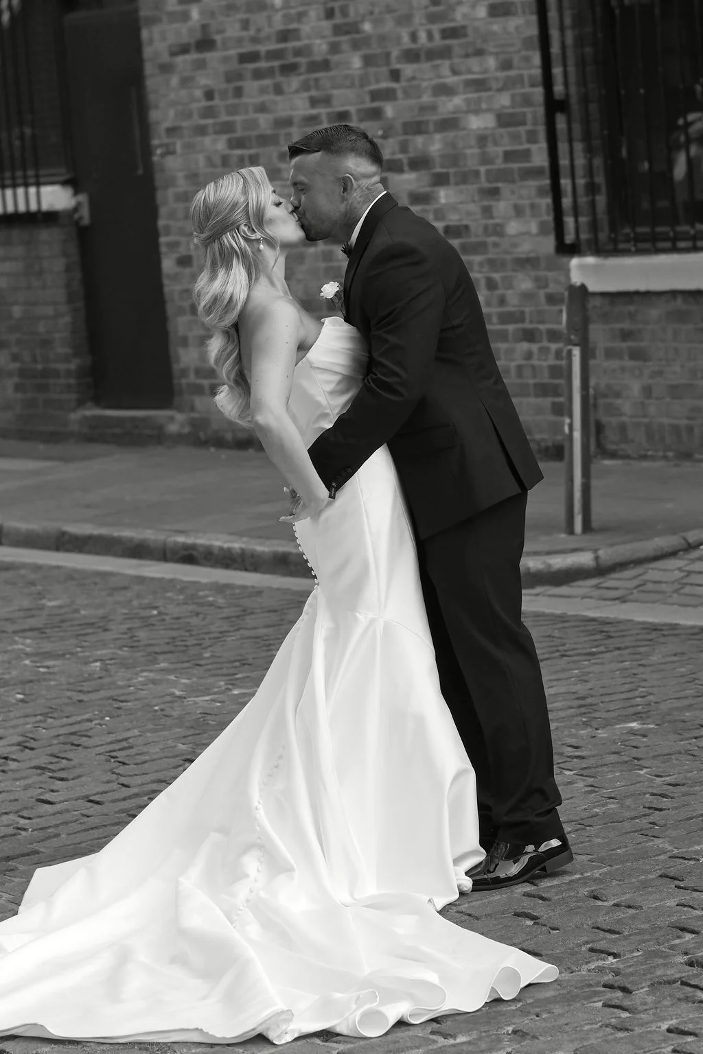 A newlywed couple share a romantic dip kiss on a cobbled city street outside an exposed brick building.