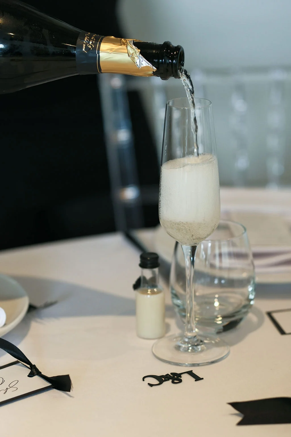 A bottle of champagne is poured into a tall flute glass set on a white linen table with place settings during the wedding reception.