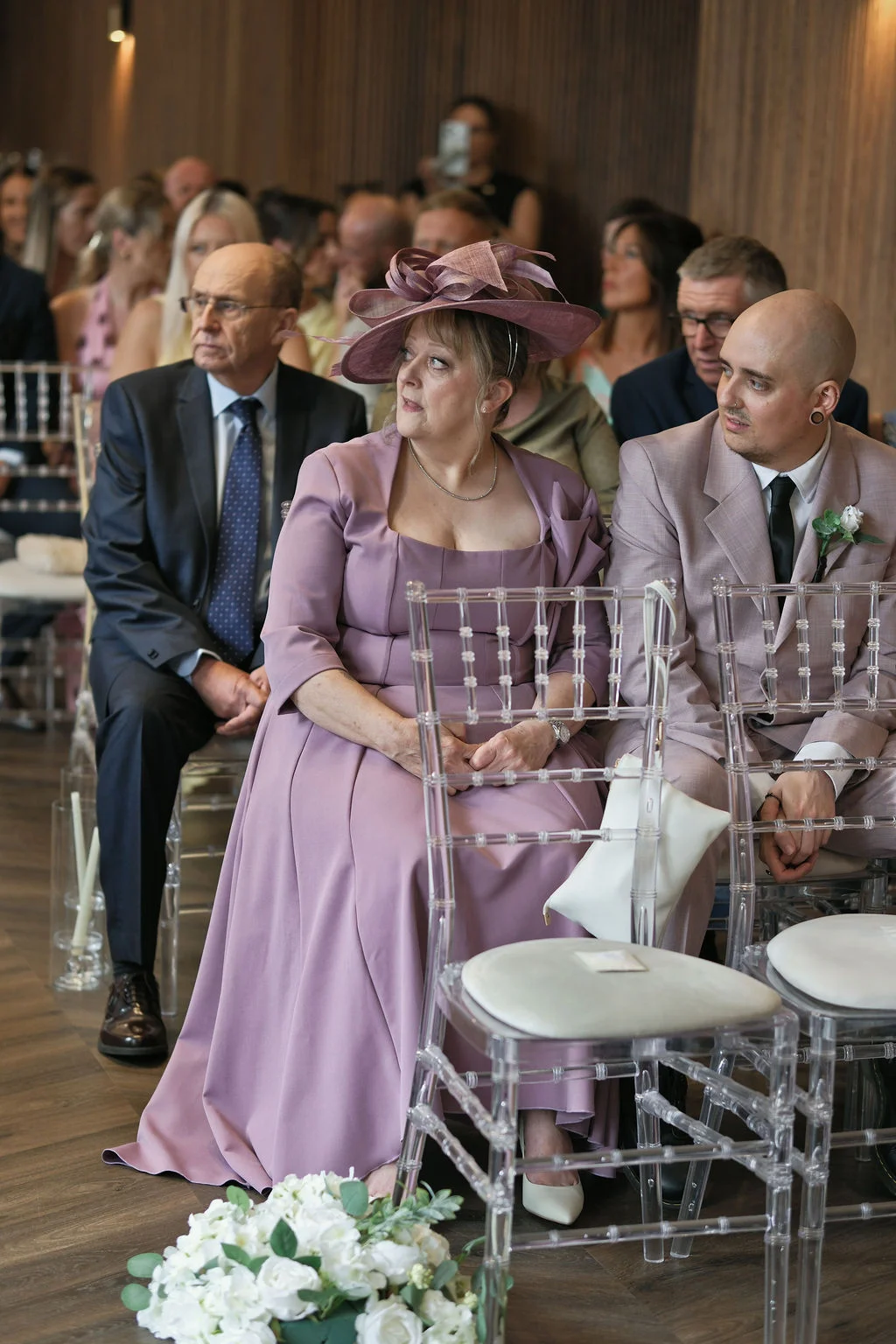 A seated wedding guest wearing a pink dress and matching fascinator looks towards the aisle while seated next to a guest in a pale pink suit during the ceremony.