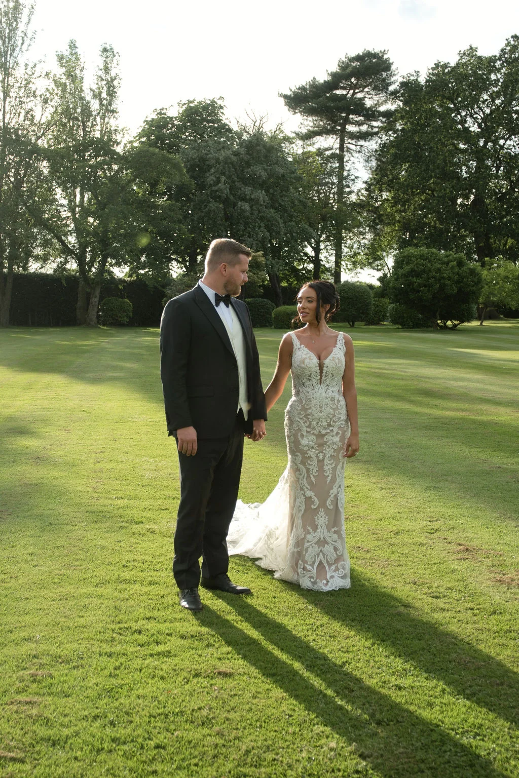 A newlywed couple holds hands while standing on a lush green lawn during golden hour, with the groom in a classic tuxedo looking lovingly at the bride in her intricately patterned lace gown.