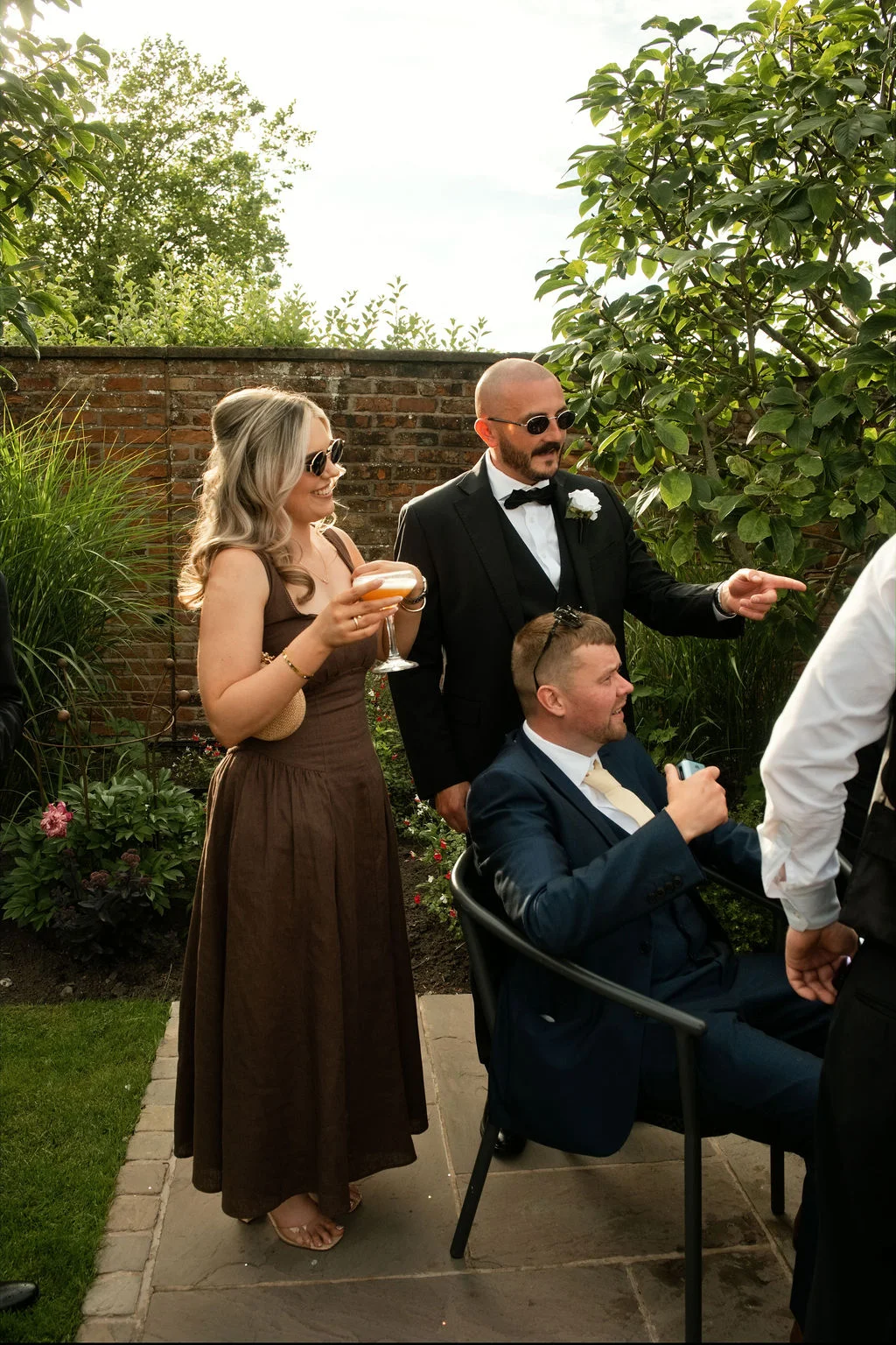 Wedding guests enjoy a drinks reception outdoors on a sunny day, with a guest in a brown dress holding a cocktail and a man in a black tuxedo looking on.