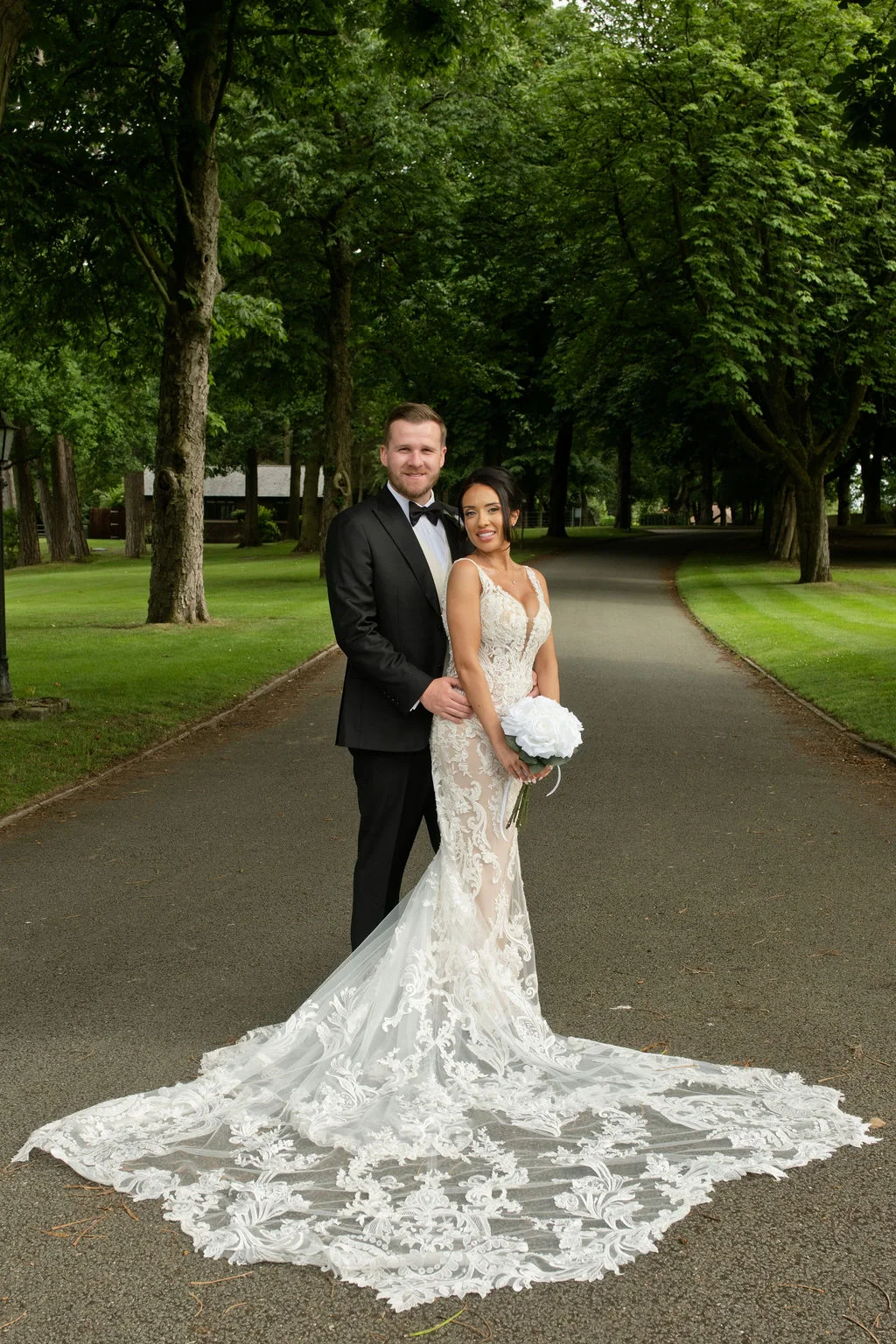 A newlywed couple poses on a paved driveway flanked by tall trees, with the bride showcasing an intricate lace wedding gown featuring a dramatic long train and holding a white bouquet.