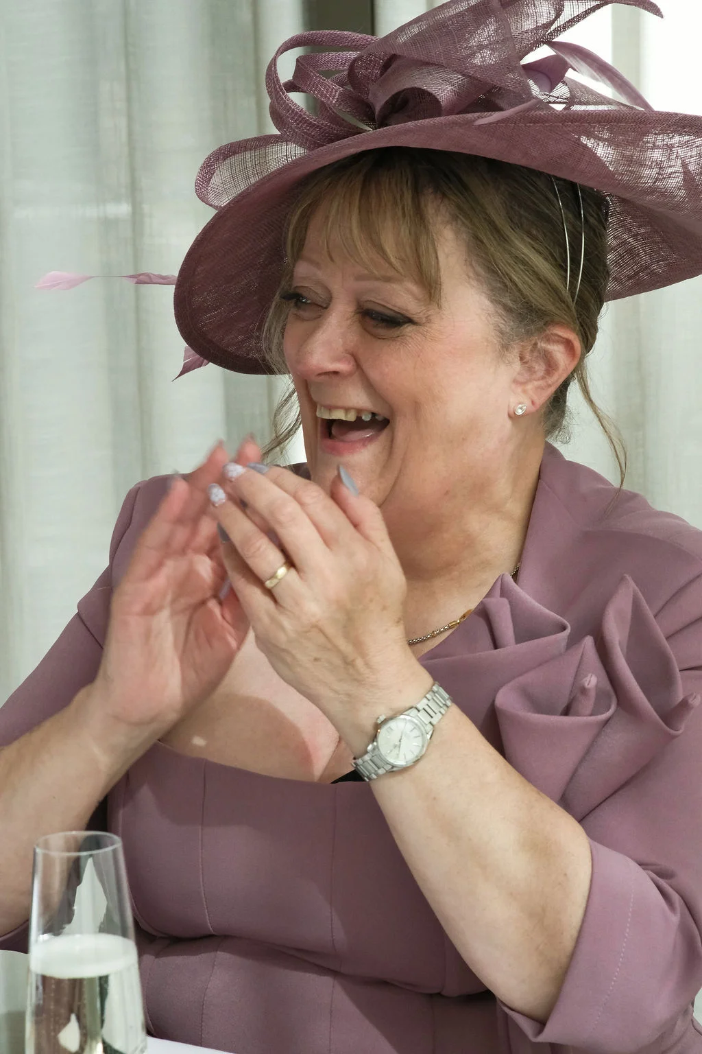 A smiling mother of the bride clapping happily while wearing a sophisticated mauve dress and matching fascinator hat at the wedding reception.