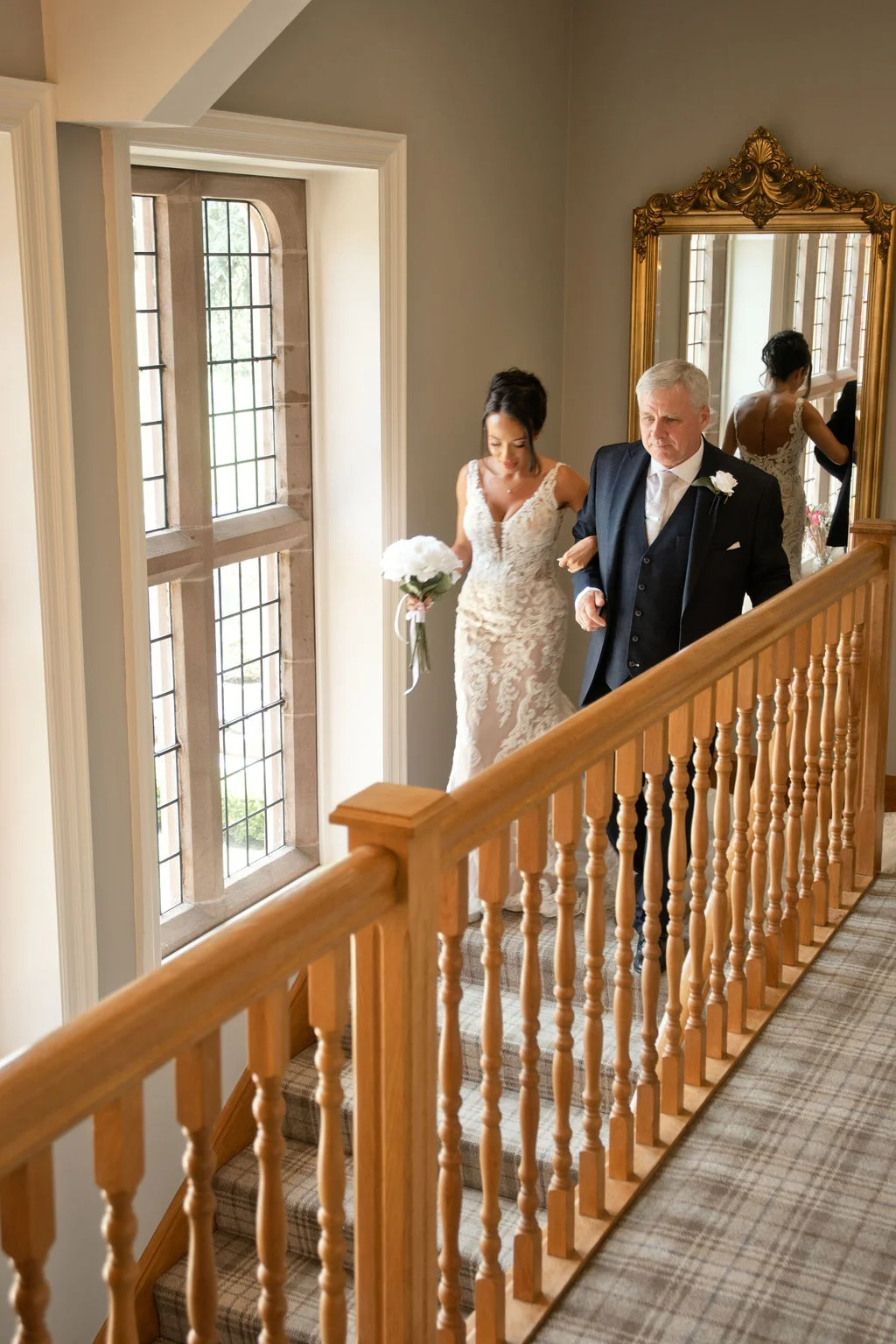 A bride in a lace sheath dress holds a white bouquet while walking down a wooden staircase alongside her father in a dark suit inside a manor house.