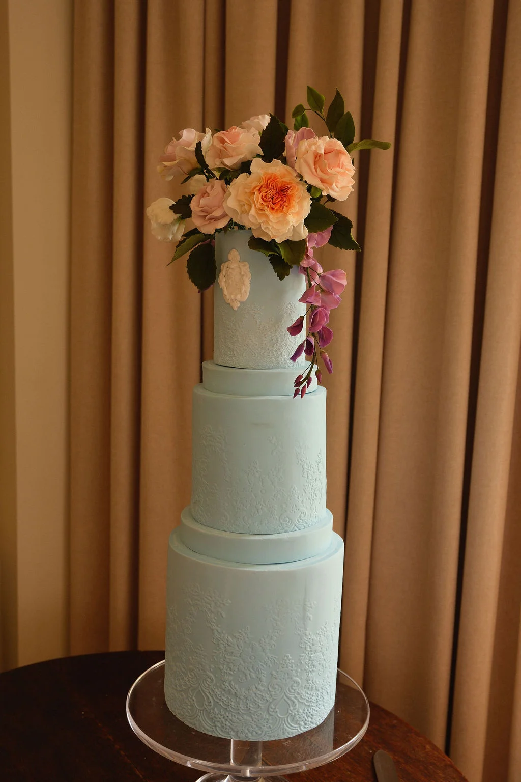A stunning three-tier pastel blue wedding cake decorated with delicate lace patterns and topped with an elaborate arrangement of peach and pink sugar roses.
