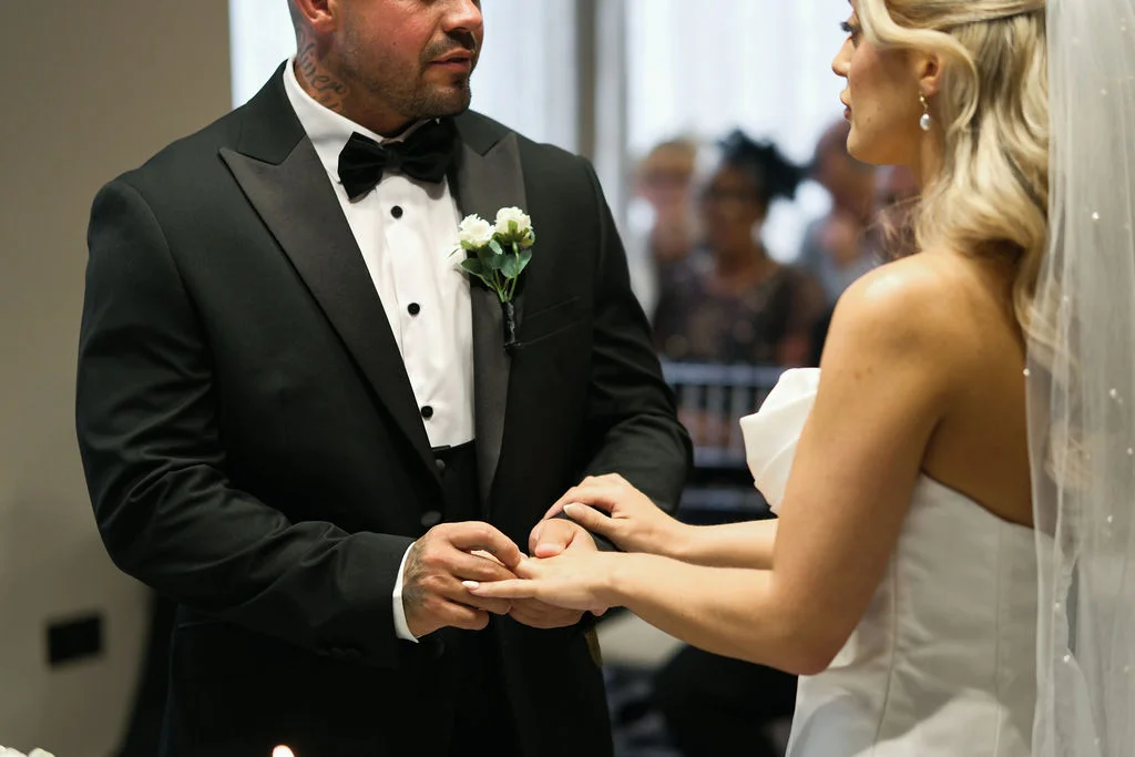 A groom in a classic black tuxedo gently places a ring on the finger of his bride during their wedding ceremony.