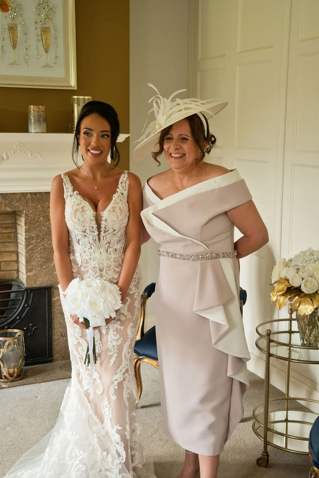 A smiling bride in a plunging lace sheath dress holding a white rose bouquet stands beside her mother wearing an elegant taupe dress and feathered fascinator indoors.