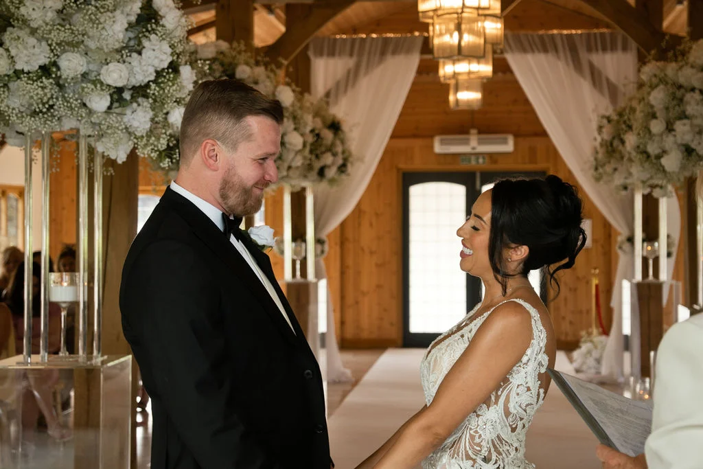 A smiling bride and groom stand face to face exchanging vows inside an oak-framed barn decorated with white floral arrangements and draping fabric.