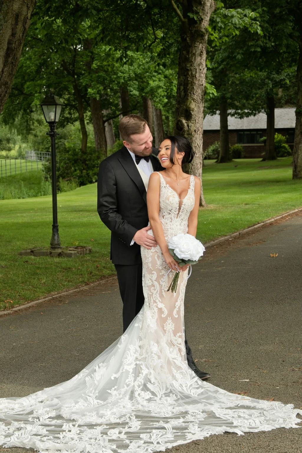 A newlywed couple embraces along a tree lined pathway, the bride holding a white rose bouquet while wearing a lace fit and flare gown with a dramatic train.