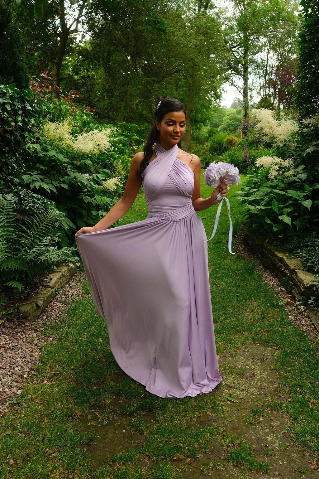 A bridesmaid in a lilac halterneck dress holds a matching bouquet while walking along a lush garden path.
