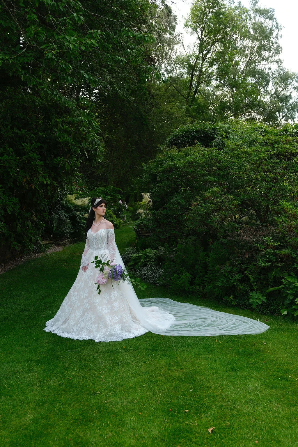 A bride in an off-the-shoulder lace gown with a long train and veil stands gracefully in a lush woodland garden, holding a bouquet of purple and pink hydrangeas.