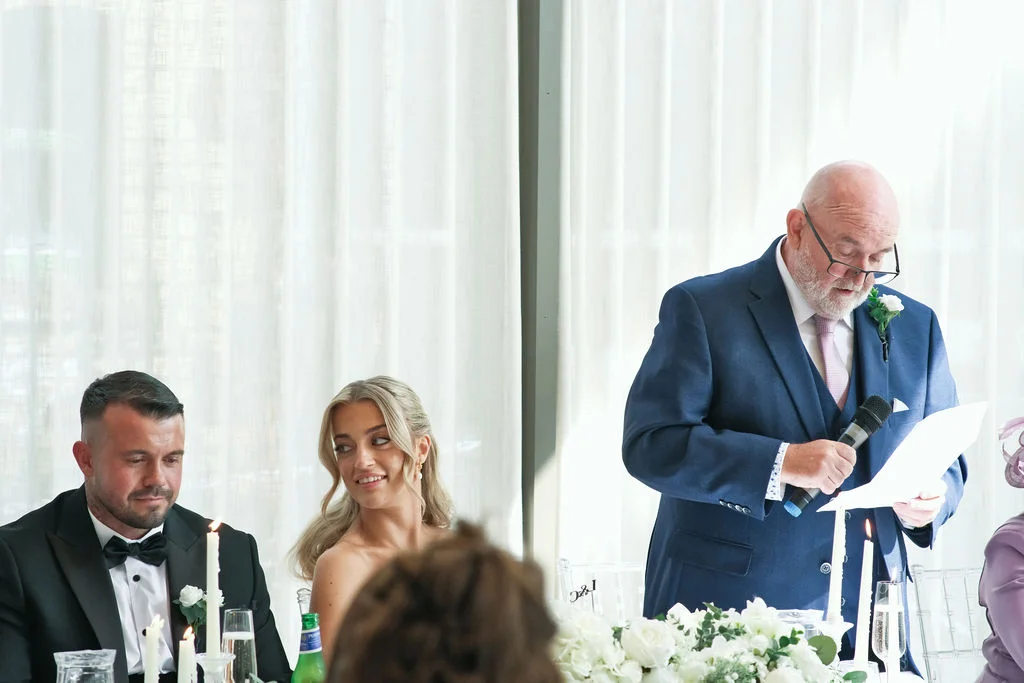 An older gentleman in a navy suit reads a speech from a paper at the wedding top table, while the smiling bride and groom look on attentively.