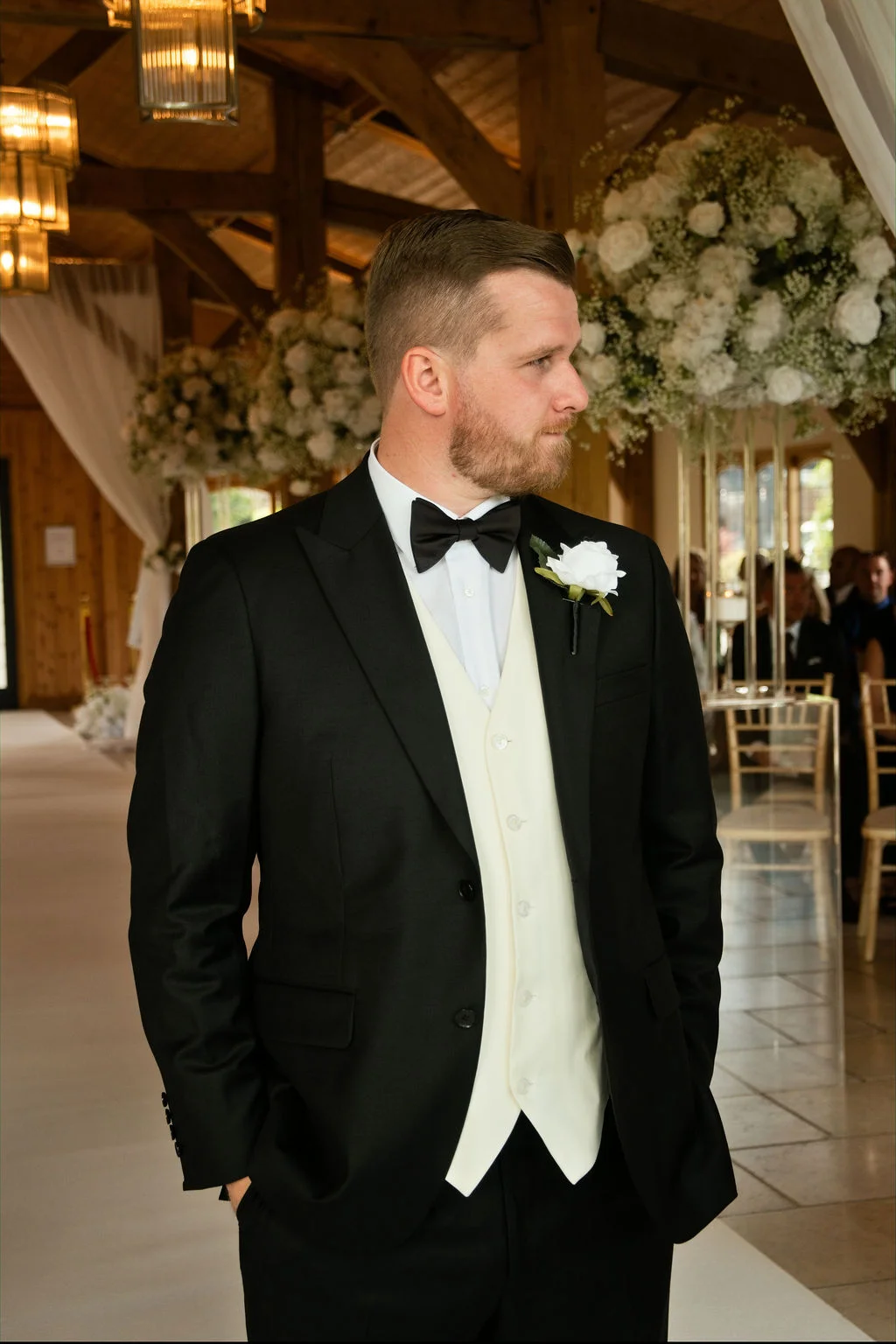 A handsome groom in a classic black suit and ivory waistcoat stands proudly inside a rustic timber-framed barn venue, wearing a white rose buttonhole as he awaits the ceremony.
